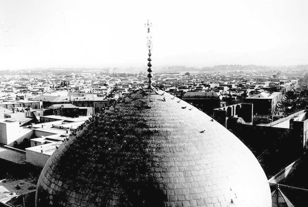 A panoramic evening view of the Imam Reza shrine in Mashhad, Iran, showing the illuminated golden dome and minarets surrounded by vast courtyards filled with pilgrims.