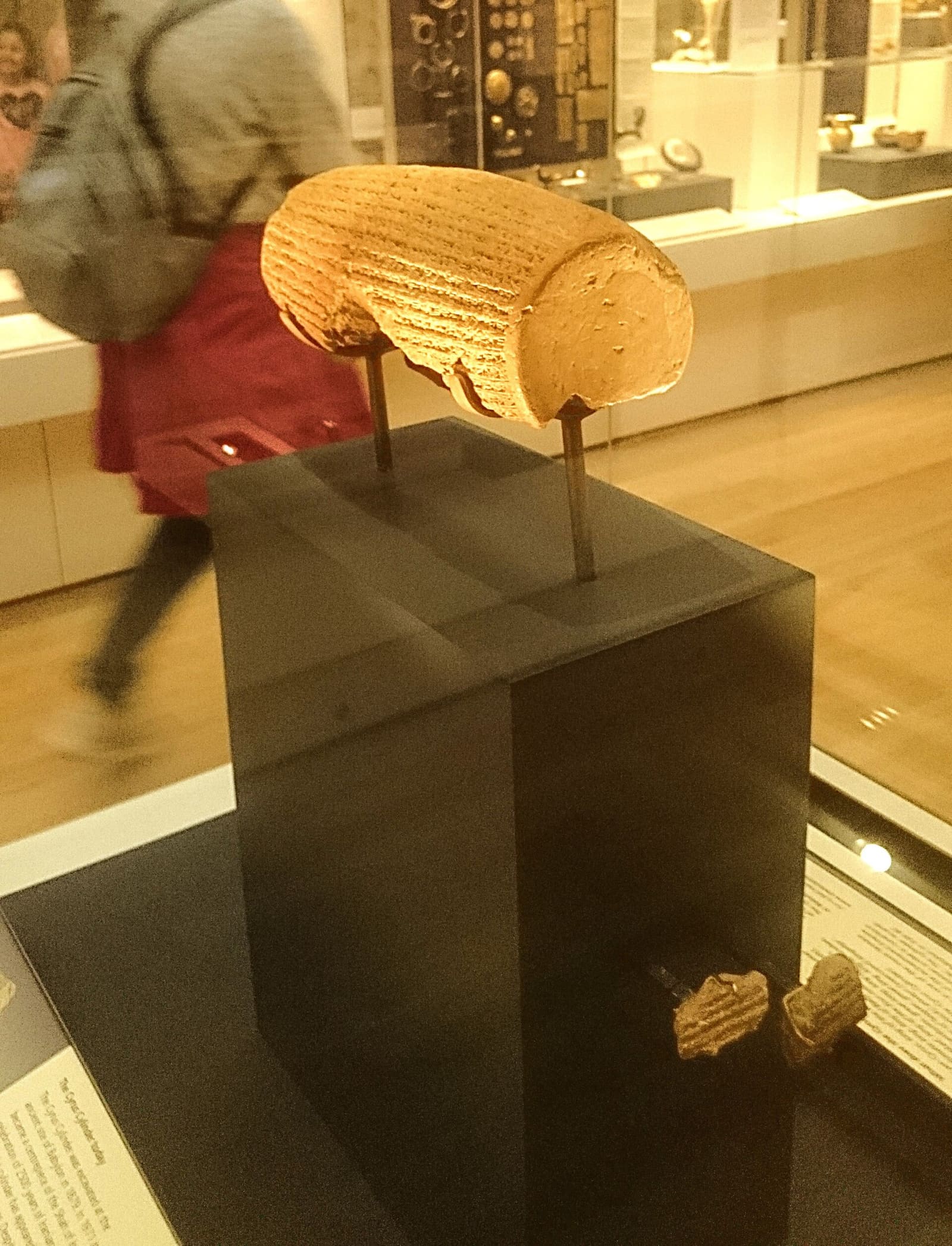 Eine detaillierte Fotografie des Kyros-Zylinders, einem tonnenförmigen Artefakt aus gebranntem Ton, das dicht mit akkadischer Keilschrift bedeckt ist und auf einer modernen Halterung ruht.