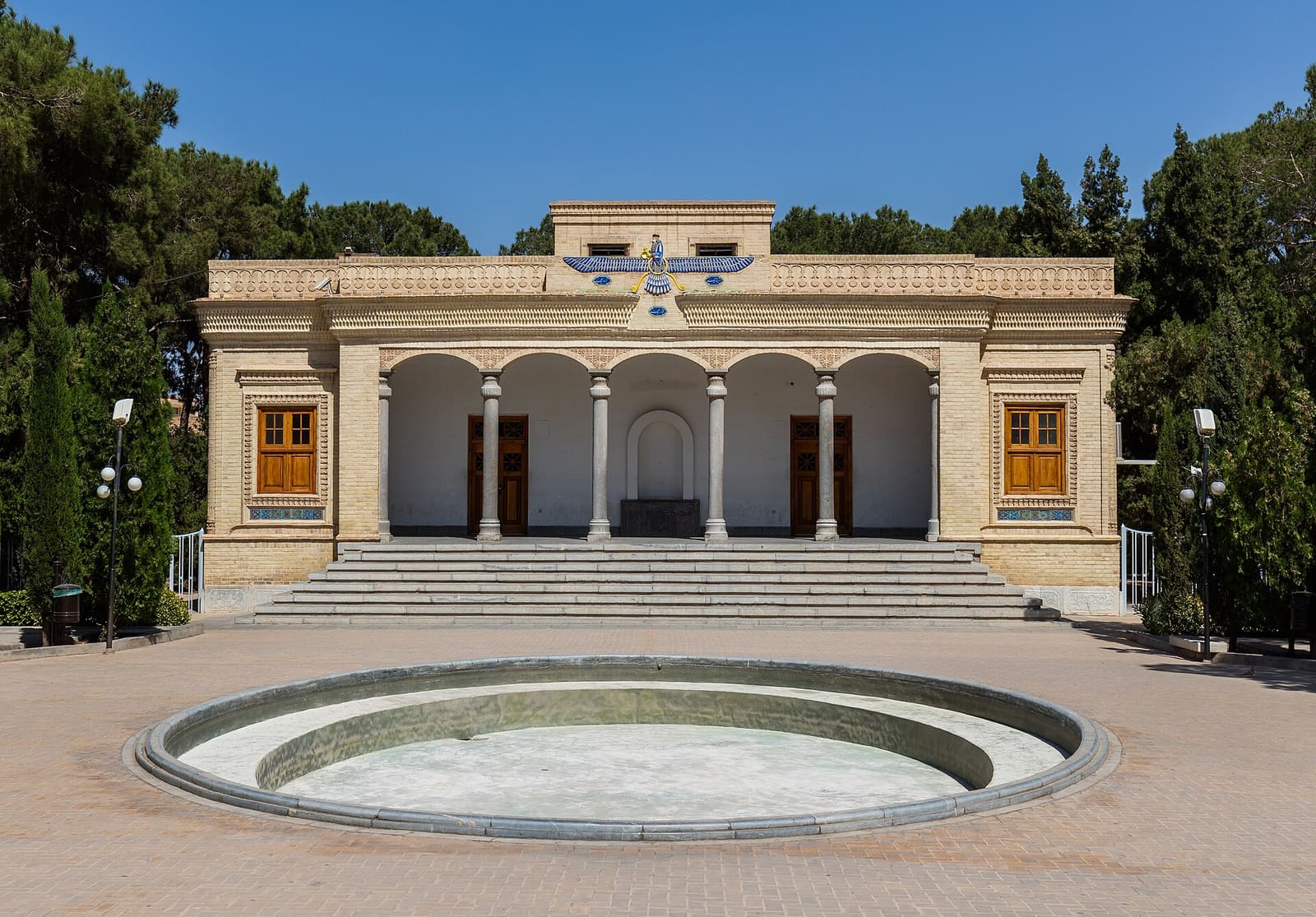 نمای بیرونی آتشکده زرتشتیان در یزد، ایران، با حوضی بزرگ در مقابل و نماد فروهر در بالای ورودی اصلی. ساختمان در روز روشن دیده می‌شود.